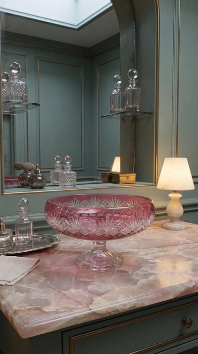 Coronet Orchid Rosé pedestal bowl centered on a blue vanity with bottles and a shaded lamp