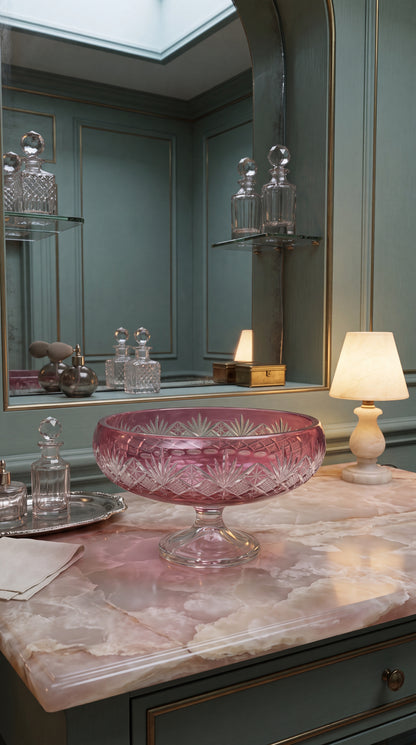 Coronet Orchid Rosé pedestal bowl centered on a blue vanity with bottles and a shaded lamp
